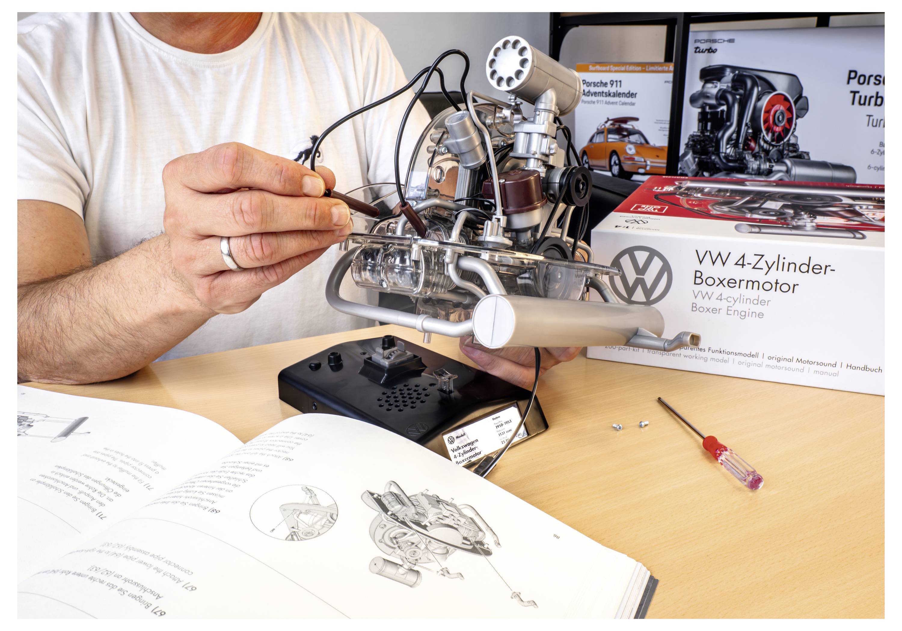 Eine Person montiert ein VW 4-Zylinder-Boxermotor-Modell mithilfe einer Anleitung. Kartonverpackung und kleine Werkzeuge sind auf dem Tisch zu sehen.