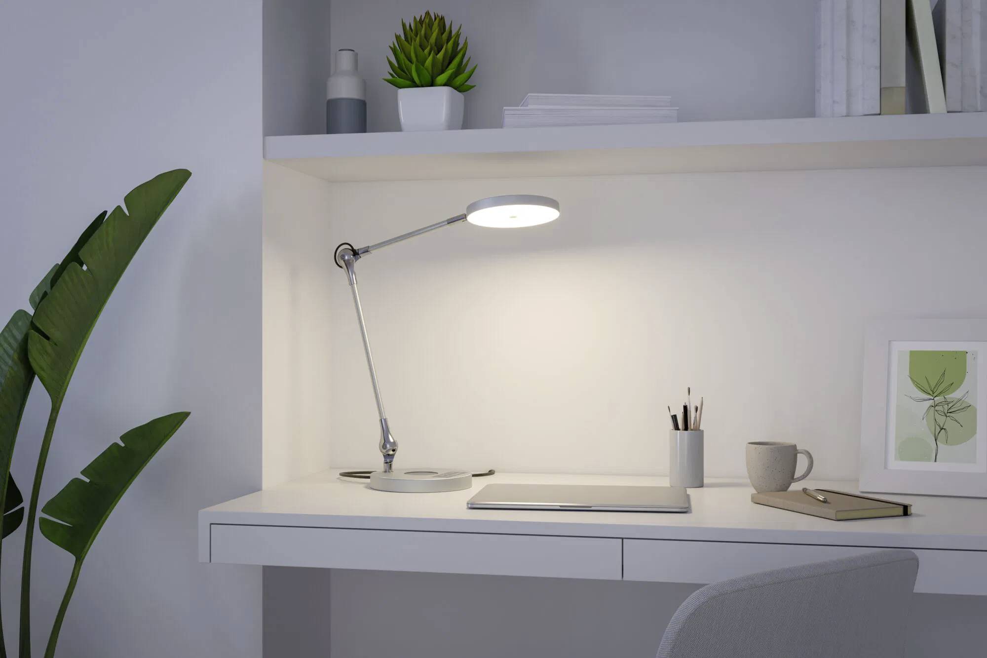 Eine minimalistische Schreibtischeinrichtung mit einer Lampe, die einen Laptop, Notizbücher und eine Tasse beleuchtet, flankiert von Pflanzen und einem gerahmten Bild auf weißen Regalen.