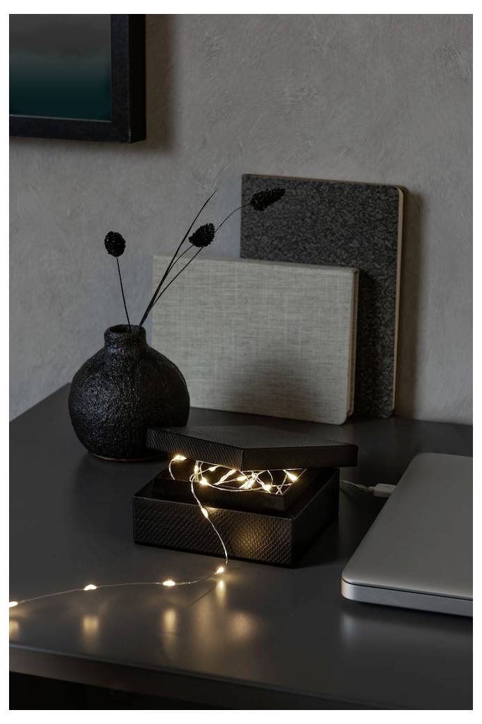Ein Schreibtisch mit einem Laptop, einer geöffneten Schachtel mit Lichterketten, zwei geschlossenen Büchern und einer strukturierten Vase mit getrockneten Pflanzen.