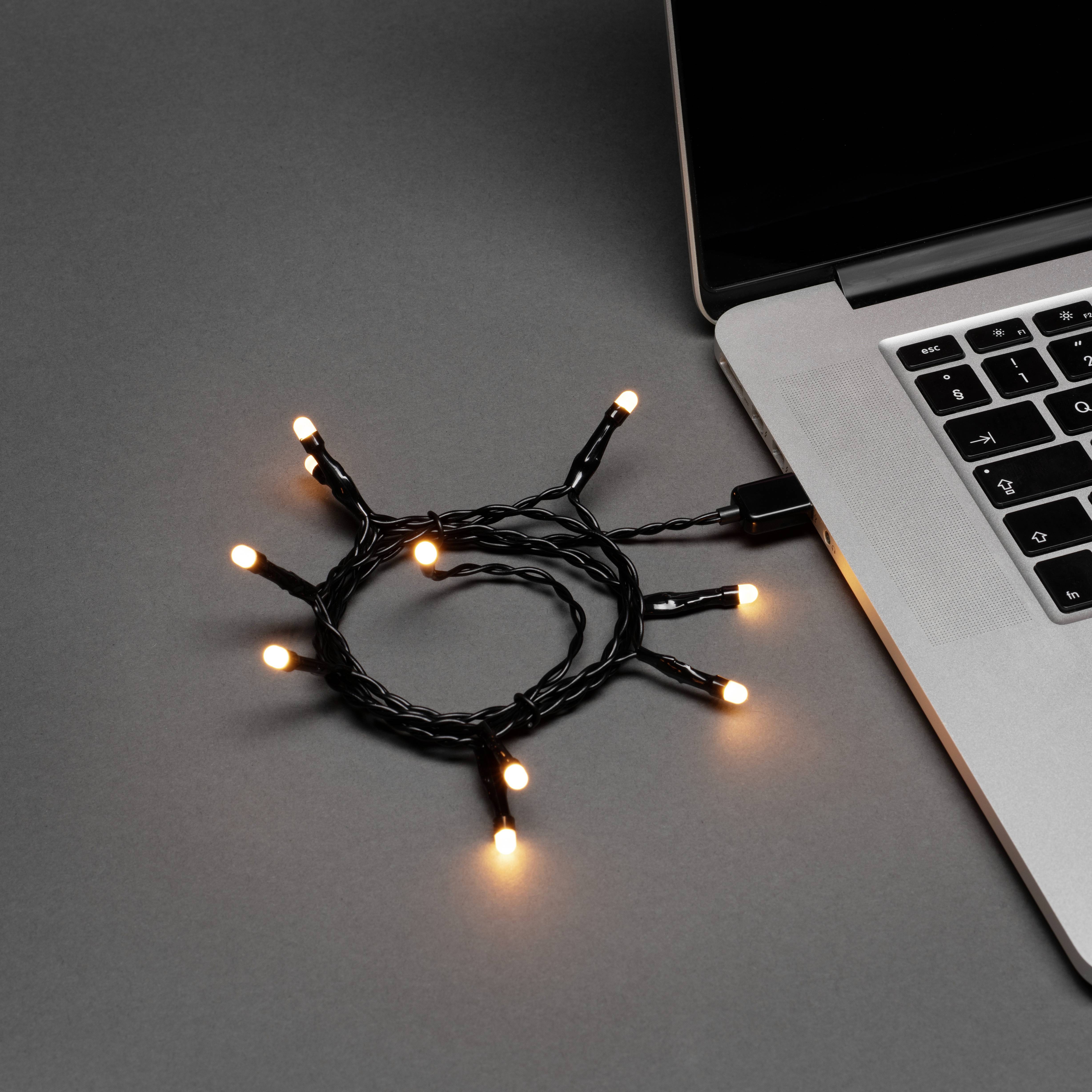 Eine Reihe von gelben Lichterketten, die in einen Laptop eingesteckt sind und vor einem grauen Hintergrund teilweise sichtbar sind, deuten auf Dekoration oder Ambiente hin.