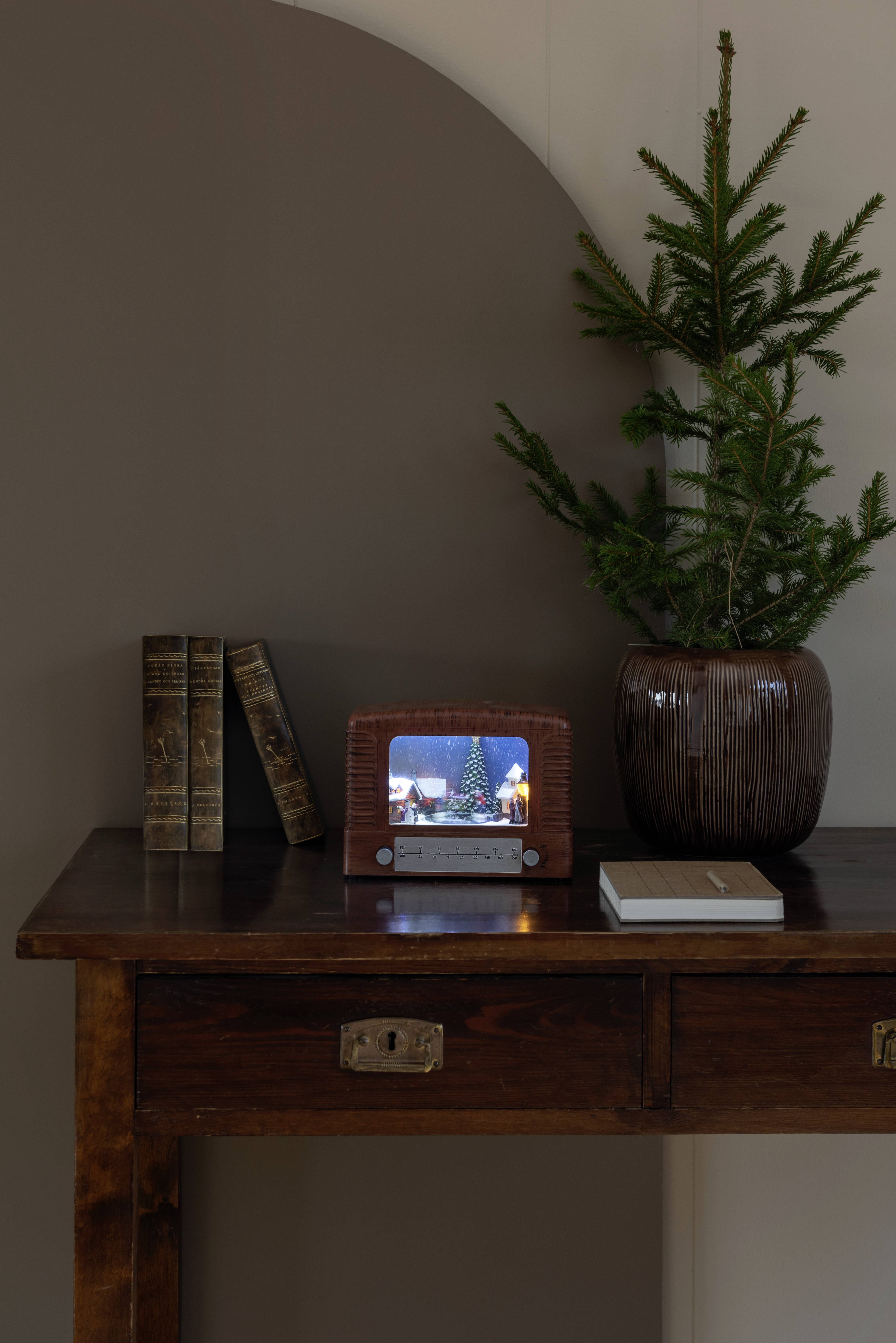 Ein kleiner Retro-Fernseher auf einem Holztisch zeigt eine Feiertagsszene mit einem Weihnachtsbaum. Eine Topfpflanze mit Nadelbäumen und Bücher stehen daneben.
