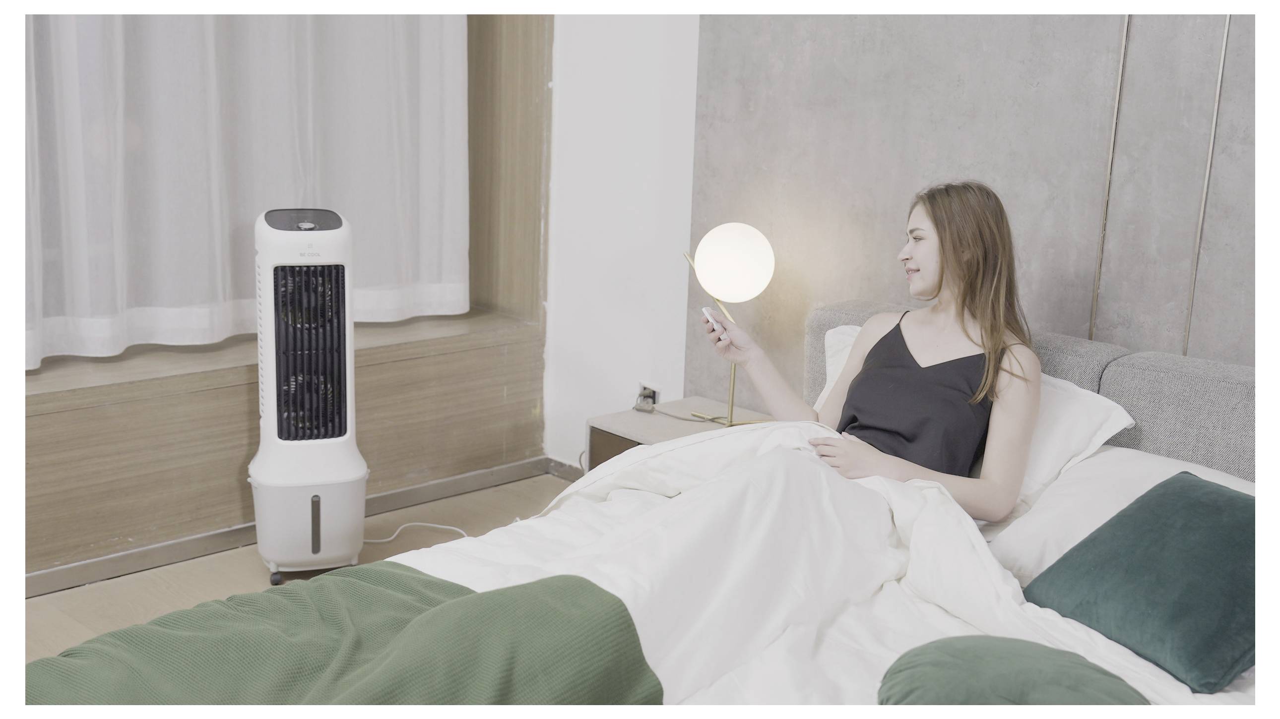 Eine Frau im Bett benutzt eine Fernbedienung, um einen Turmventilator zu bedienen, der in der Nähe eines Fensters steht.