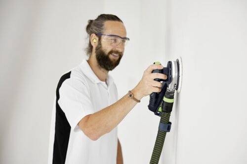 Eine Person mit Sicherheitsbrille und Ohrstöpseln benutzt einen elektrischen Schleifer an einer weißen Wand und demonstriert eine Schleiftechnik.