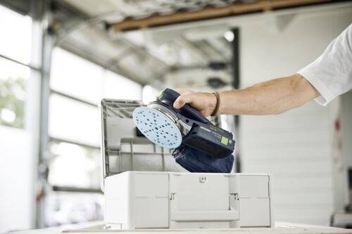 Eine Person legt einen Exzenterschleifer in eine Aufbewahrungsbox in einer hell erleuchteten Werkstatt. Der Deckel der Box ist geöffnet und zeigt einen ordentlich arrangierten Innenraum.