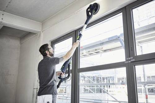 Eine Person verwendet eine Exzenterschleifer, um eine Wand in der Nähe eines großen Fensters in einem Bau- oder Renovierungsumfeld zu glätten.