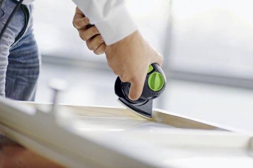 Eine Person schleift eine Holzoberfläche mit einem tragbaren elektrischen Schleifgerät und konzentriert sich darauf, die Kante zu glätten. Die Umgebung ist ein heller, moderner Arbeitsbereich.