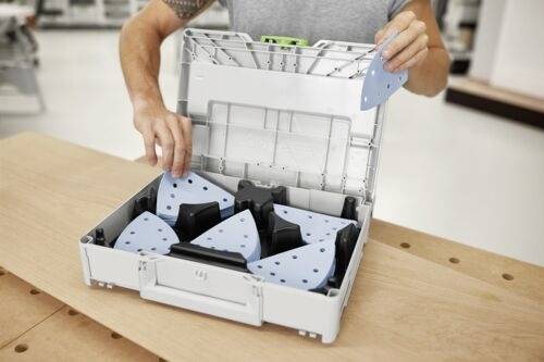 Eine Person ordnet dreieckige Schleifblätter in einer Plastikaufbewahrungsbox auf einer Holzwerkbank an, bereit für eine Schleifaufgabe.
