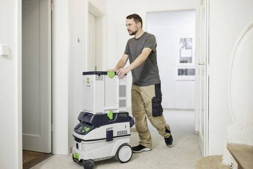 Eine Person schiebt eine große mobile Filtereinheit durch einen hellen Flur, was auf den Einsatz der Ausrüstung zur Luftreinigung oder Reinigungsaufgaben hindeutet.