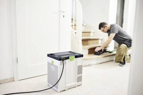 Ein Mann schleift eine Holztreppe mit einem elektrischen Schleifgerät. Ein Staubsauger steht in der Nähe und ist über einen Schlauch mit dem Schleifgerät verbunden.