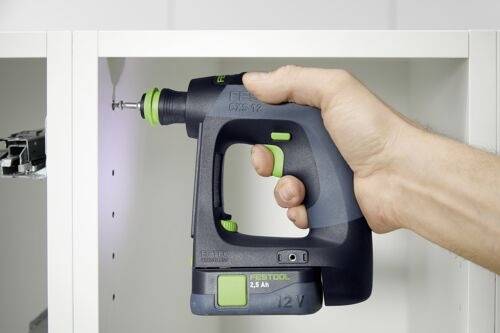 Eine Person verwendet einen kabellosen Akkuschrauber, um eine Schraube in einem weißen Schrank festzuziehen. Der Akkuschrauber ist schwarz mit grünen Akzenten und trägt die Aufschrift ‚FESTOOL 12V'.