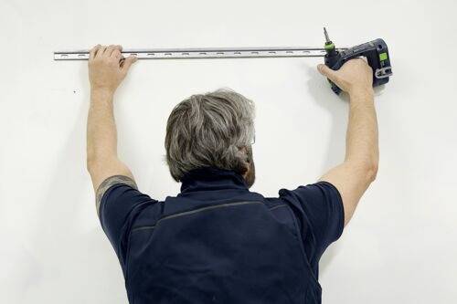 Eine Person misst eine weiße Wand mit einem Maßband und hält eine Bohrmaschine, um etwas zu montieren oder zu installieren.