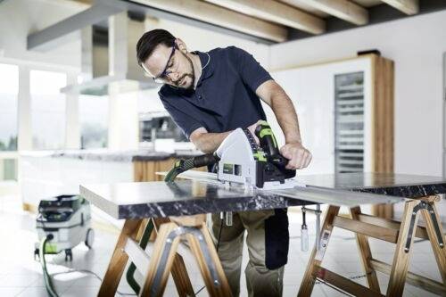 Eine Person, die eine Tauchsäge verwendet, um ein Stück Holz drinnen zu schneiden, trägt Schutzbrille und steht zwischen zwei Böcken in einer modernen Küche.
