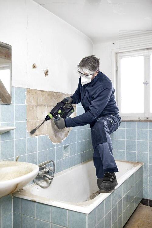Eine Person in Schutzausrüstung renoviert ein Badezimmer und verwendet ein Elektrowerkzeug, um Fliesen von einer Wand über einer Badewanne zu entfernen.