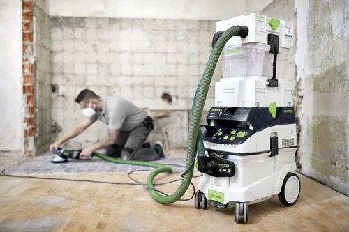 Eine Person mit Maske schleift einen Holzboden in einem teilweise renovierten Raum. Ein Staubabsauggerät ist prominent im Vordergrund.