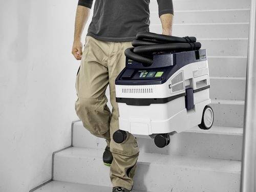 Eine Person, die einen tragbaren Staubsauger eine Treppe hinunterträgt und dabei sein geringes Gewicht und seine benutzerfreundliche Handhabung demonstriert.