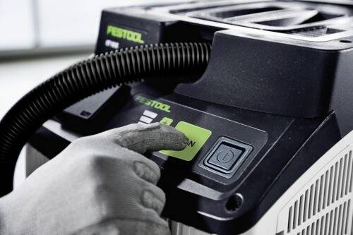 Eine Person bedient einen Festool Staubsauger und drückt dabei den grünen 'EIN'-Knopf. Das Gerät verfügt über einen spiralförmigen Schlauch und eine Steuerungsoberfläche.