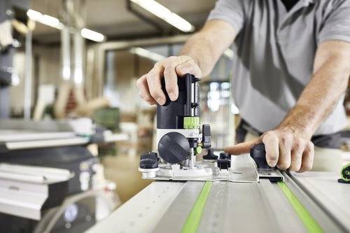 Eine Person bedient eine Tischfräse in einer Werkstatt und führt Holz durch die Maschine. Der Arbeitsbereich ist gut beleuchtet, mit verschiedenen Werkzeugen im Sichtfeld.