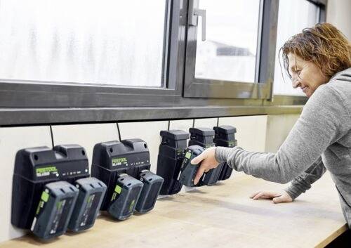 Eine Person legt eine Batterie in ein Ladegerät auf einer Werkbank mit mehreren Ladegeräten und Batterien. Ein großes Fenster sorgt für natürliches Licht.