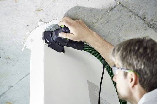Eine Person mit Sicherheitsbrille benutzt ein Elektrowerkzeug, um eine Oberfläche zu schleifen oder zu polieren, wobei ein grüner Schlauch zur Staubabsaugung angeschlossen ist.
