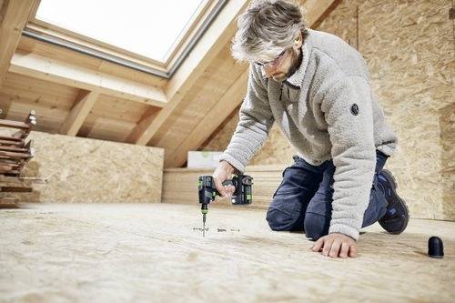 Eine Person kniet auf einem Holzboden und installiert Schrauben mit einer Elektrobohrmaschine in einem hellen Dachbodenraum.