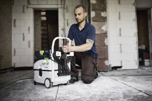 Eine Person kniet auf einem Baustellenboden und justiert eine kompakte Reinigungsmaschine mit Griff und Rädern. Der Hintergrund zeigt unfertige Wände.