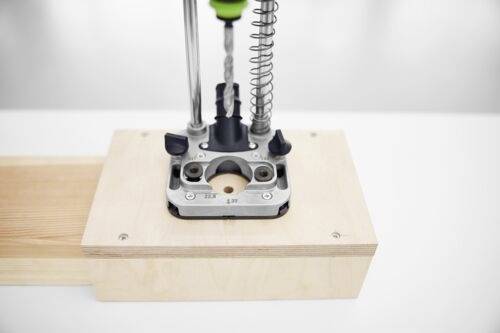 Eine Bohrmaschine, die auf einer Holzplattform positioniert ist und einen Metallbohrer mit Federmechanismus aufweist, der nach unten auf die Holzoberfläche gerichtet ist.