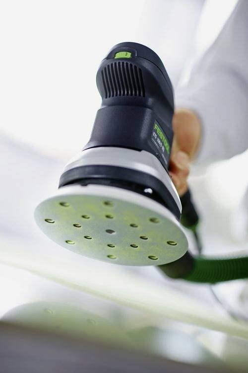 Eine Hand, die eine Exzenterschleifer mit grün-schwarzem Griff hält, von unten betrachtet, wobei der kreisförmige Schleifpad mit Löchern zu sehen ist.