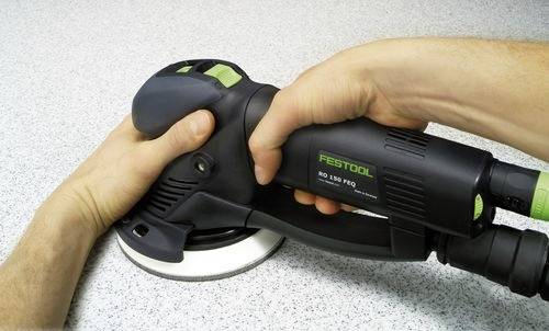 Eine Person bedient einen Festool RO 150 FEQ Schleifer auf einer hellgrauen gesprenkelten Oberfläche, wobei eine Nahaufnahme der Hände zu sehen ist, die das Werkzeug sicher umfassen.
