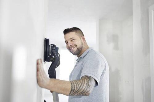 Eine Person benutzt einen Wandschleifer, um eine Wandoberfläche in einem Raum zu glätten und trägt dabei ein lässiges Hemd und zeigt einen tätowierten Arm.