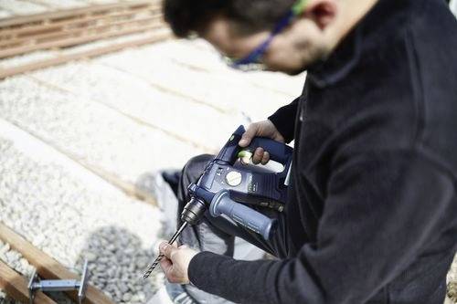 Eine Person mit Sicherheitsbrille bedient einen Bohrer auf einer Baustelle und konzentriert sich auf präzises Bohren in eine Oberfläche, die von Kies umgeben ist.
