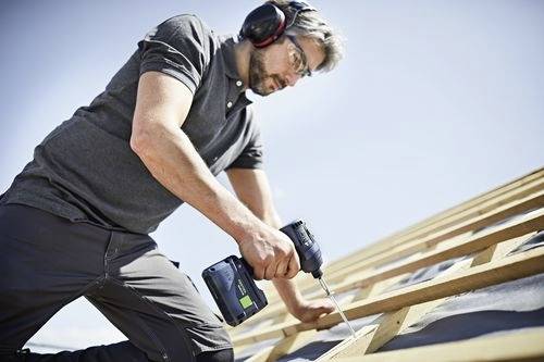 Ein Arbeiter auf einem Dach verwendet eine Bohrmaschine, um Holzbalken zu befestigen und trägt dabei Schutzausrüstung. Der klare Himmel im Hintergrund deutet auf eine Baustelle hin.
