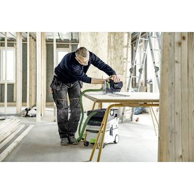 Eine Person in Arbeitskleidung bedient ein Elektrowerkzeug an einem Holztisch in einem unfertigen Raum, was auf ein laufendes Bauprojekt hindeutet.