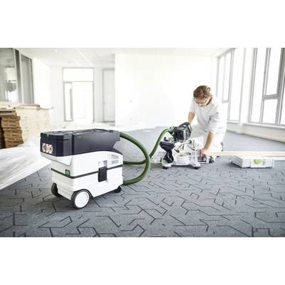 Eine Person, die ein Elektrowerkzeug verwendet, um Bodenmaterial in einem hellen, unfurnissierten Raum zu schneiden, wobei ein Staubabsauggerät in der Nähe steht.