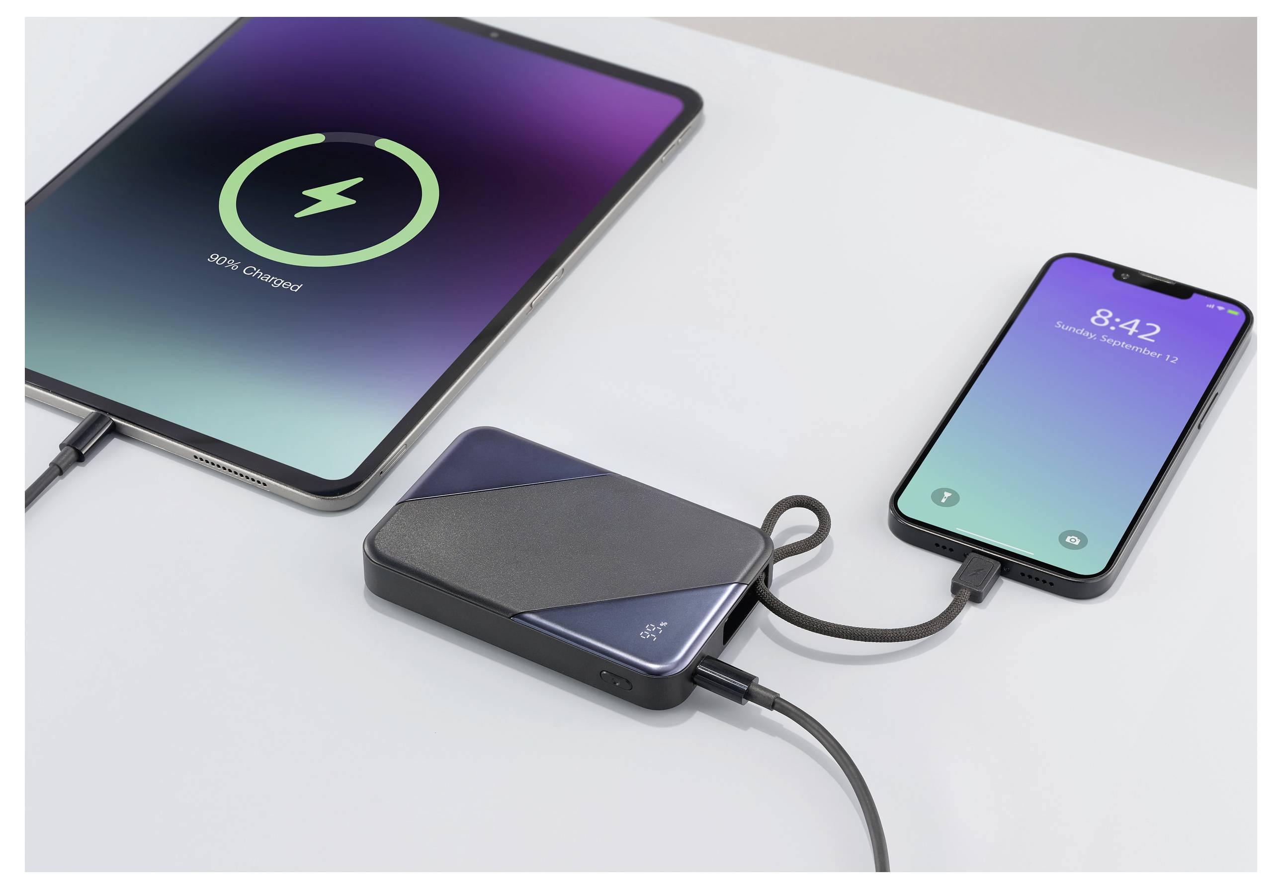 Ein Tablet und ein Smartphone sind mit einem tragbaren Ladegerät auf einem Tisch verbunden. Der Tablet-Bildschirm zeigt „58% aufgeladen" an; das Telefon zeigt „8:42" an.