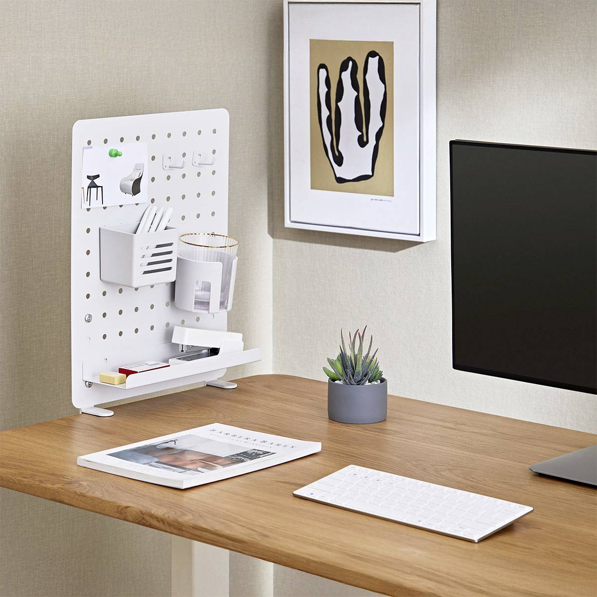 Moderner Büroschreibtisch mit Lochplatte-Organizer, Pflanze und gerahmter abstrakter Kunst an der Wand; minimalistische Arbeitsplatz-Einrichtung mit Tastatur.