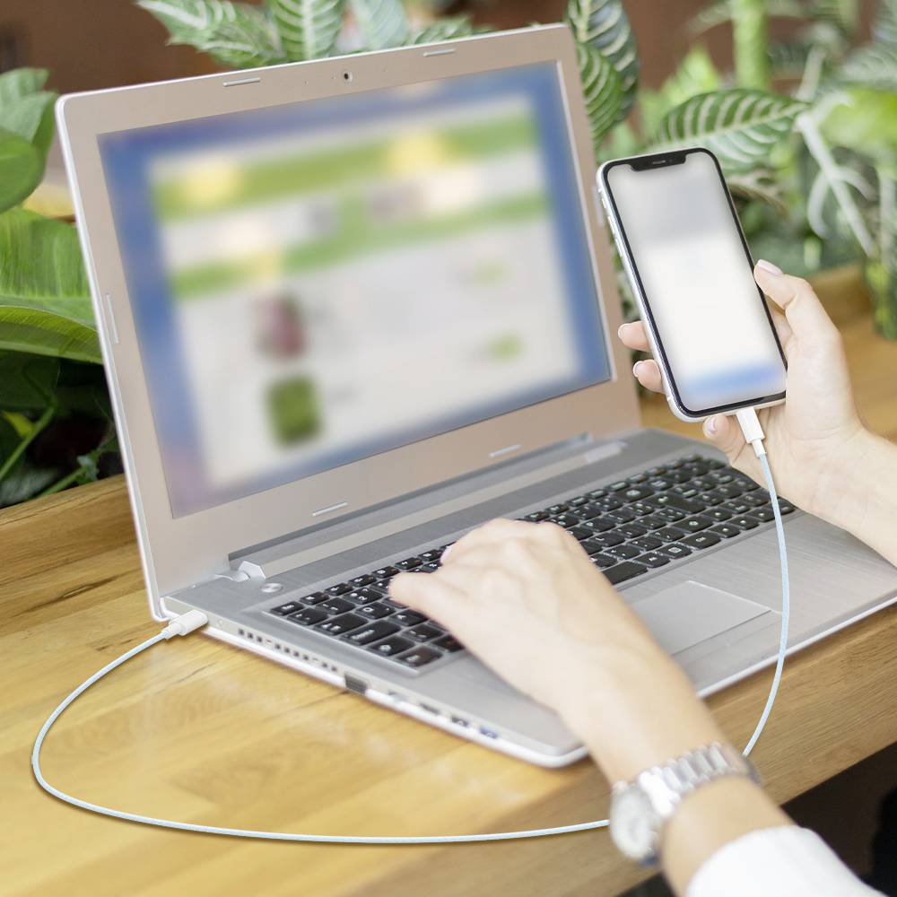 Eine Person benutzt ein Smartphone, das mit einem Laptop auf einem Holzschreibtisch verbunden ist. Der Laptop-Bildschirm zeigt eine verschwommene Website und deutet damit auf Multitasking hin.