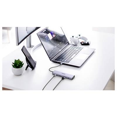 Ein Laptop, der an einer Dockingstation auf einem weißen Schreibtisch angeschlossen ist, mit einem Smartphone-Ständer, einem Gamepad und einem Monitor im Hintergrund.