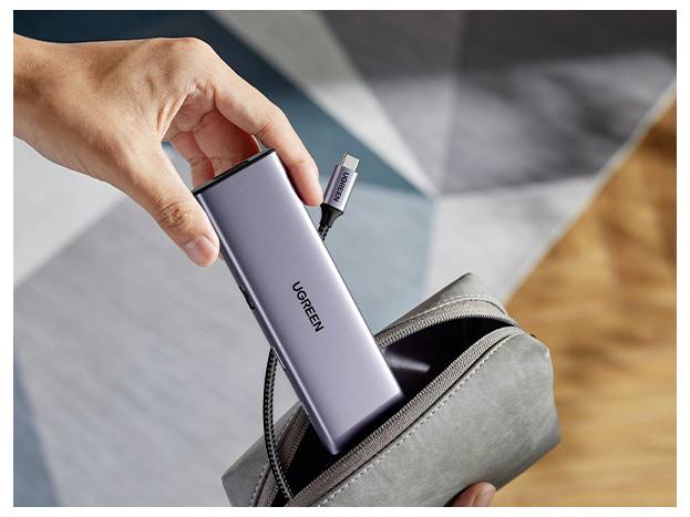 Eine Hand hält einen grauen UGREEN USB-C Hub über einen grauen Beutel, bereit zum Verstauen. Das Kabel des Hubs ist teilweise sichtbar.