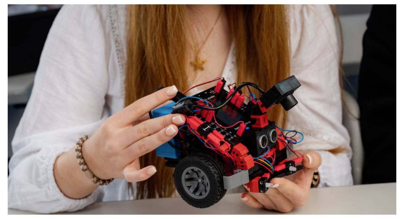 Eine Person, die ein kleines, rotes Roboterfahrzeug mit verschiedenen elektronischen Komponenten und Rädern hält und dabei ein Robotik-Projekt präsentiert.