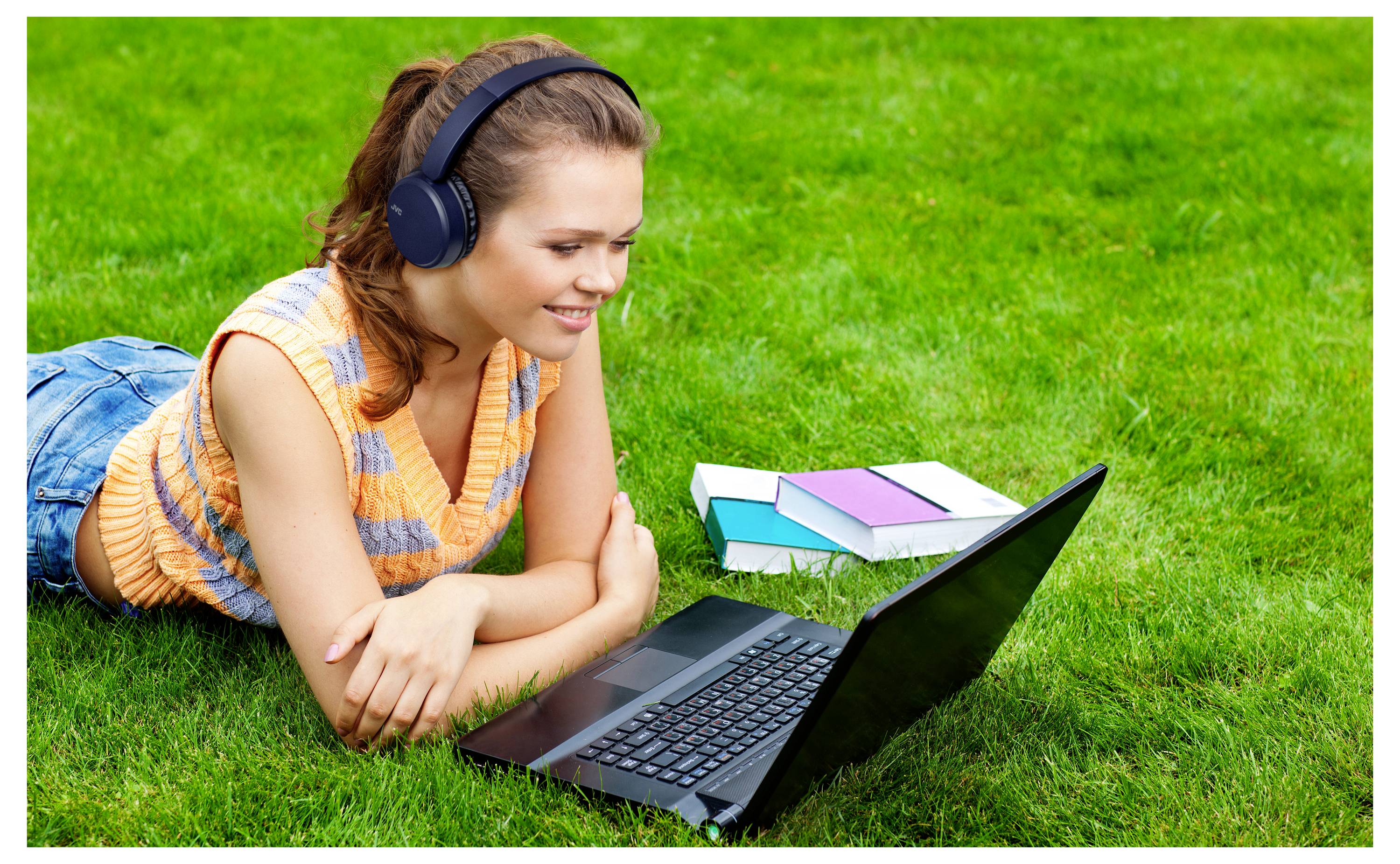 Eine Person mit Kopfhörern liegt auf dem Gras und schaut auf einen Laptop. Bücher liegen in der Nähe und deuten auf eine Studien- oder Freizeitaktivität hin.