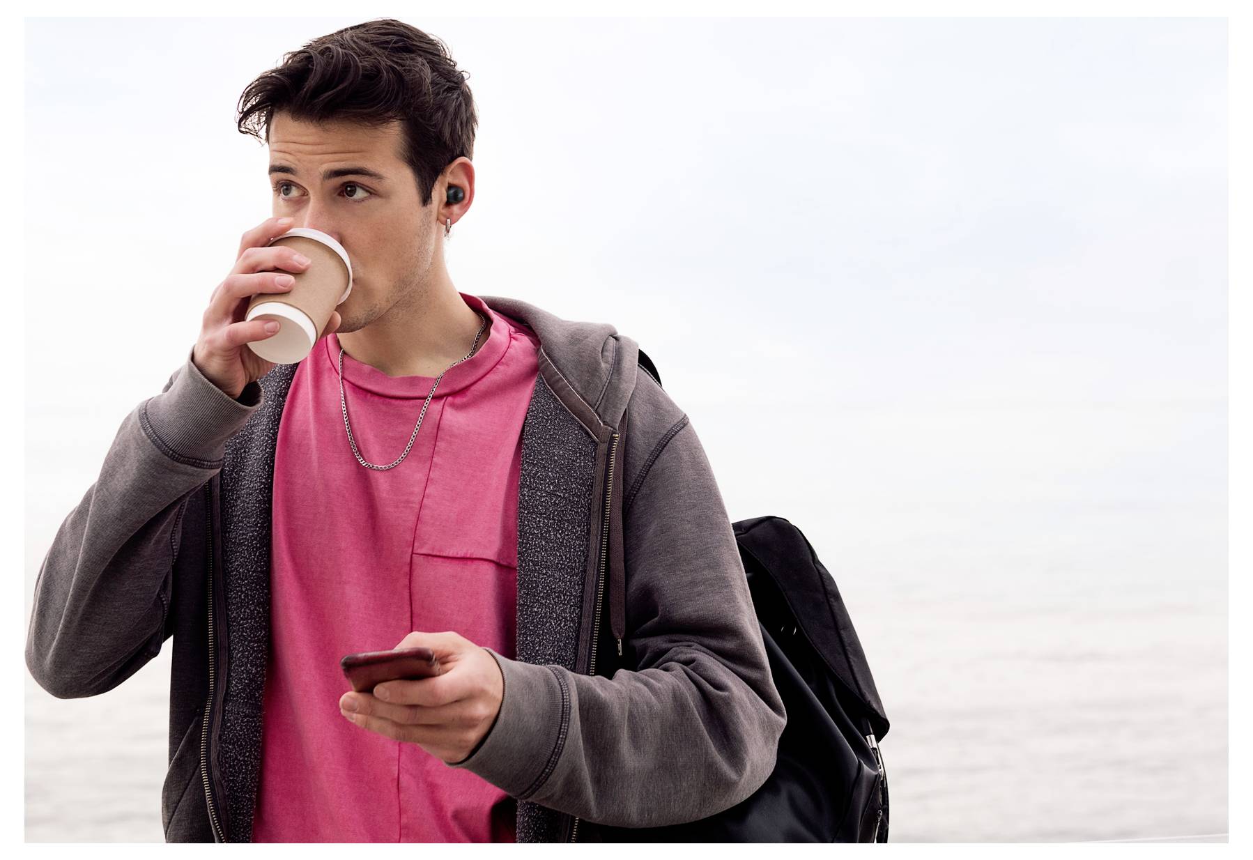 Mann in einem rosa Hemd und grauem Hoodie trinkt Kaffee und hält ein Telefon, trägt Kopfhörer, mit Meeresblick im Hintergrund.