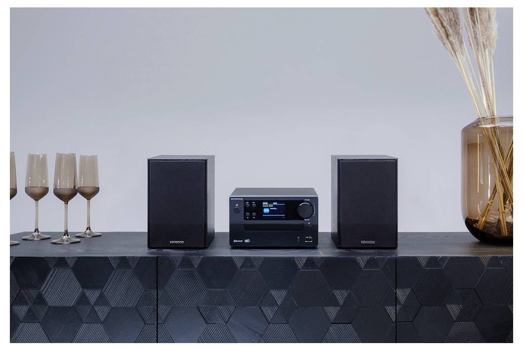 Ein schlankes Stereosystem mit zwei Lautsprechern steht auf einer geometrisch gemusterten Oberfläche, begleitet von einem Satz Gläsern und einer hohen Vase.