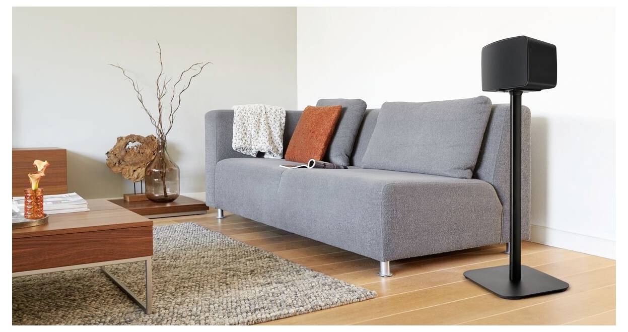 Ein modernes Wohnzimmer zeigt ein graues Sofa mit einem roten Kissen, eine Zierpflanze und einen Lautsprecher auf einem Ständer, was einen gemütlichen, technikfreundlichen Raum andeutet.