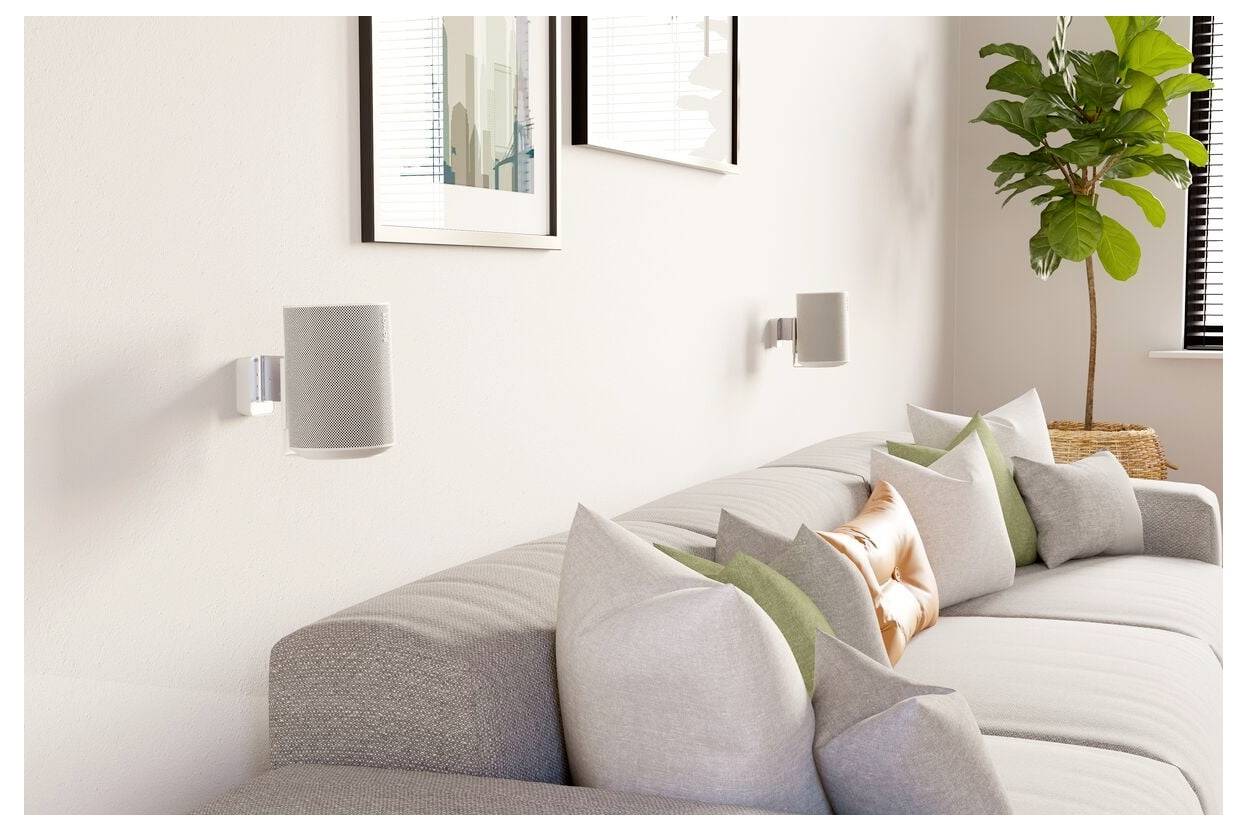 Ein modernes Wohnzimmer präsentiert ein großes, hellgefärbtes Ecksofa, das mit verschiedenen strukturierten Kissen geschmückt ist. Wandmontierte Lautsprecher und gerahmte Kunstwerke schmücken die Wände, und eine Topfpflanze verleiht dem Raum Grün.