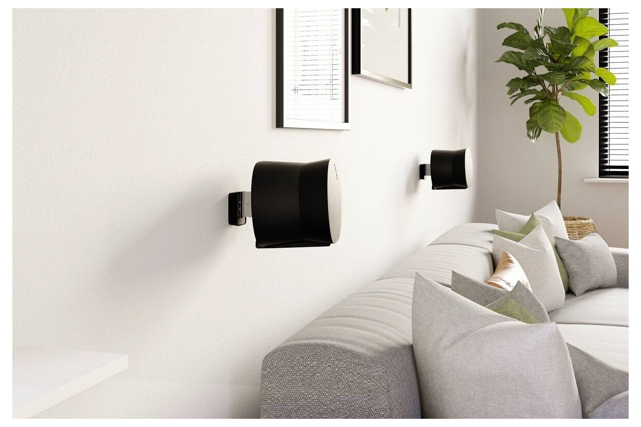 Wohnzimmer mit einem modernen grauen Sofa, zwei wandmontierten schwarzen Lautsprechern, gerahmter Kunst und einer Topfpflanze in der Nähe eines Fensters mit Jalousien.