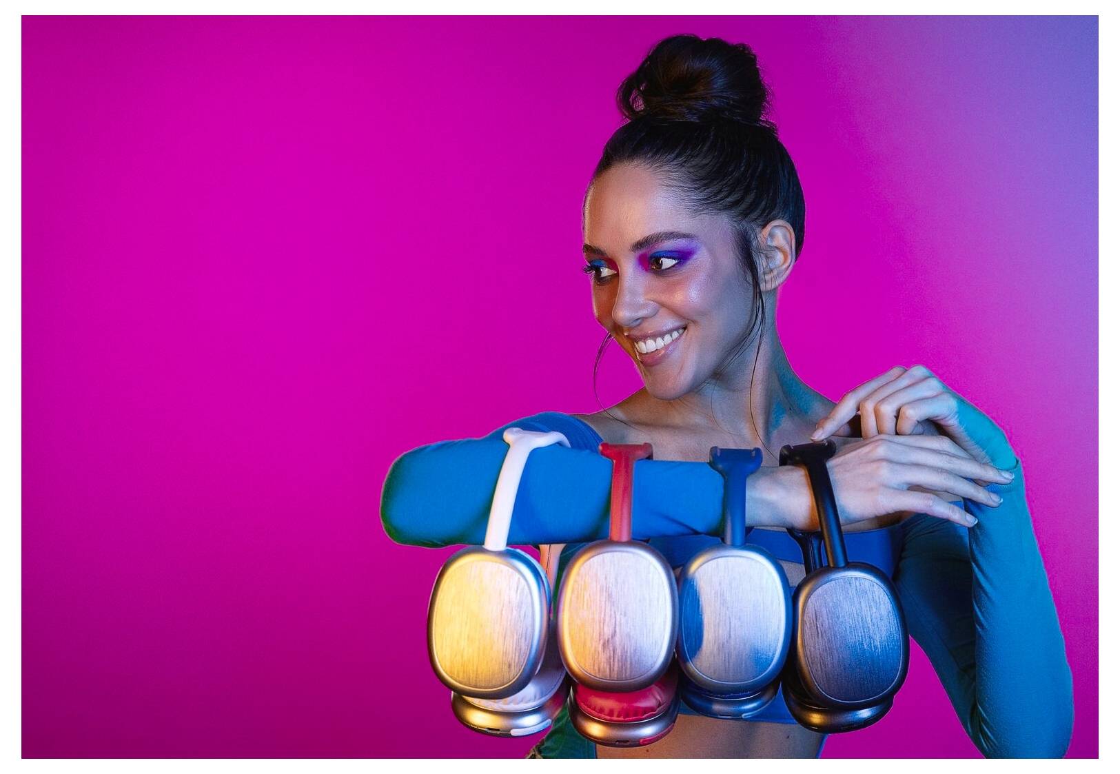 Eine Person in farbenfroher Beleuchtung hält vier Kopfhörerpaare in verschiedenen Farben und präsentiert Tech-Mode vor einem leuchtend pinken Hintergrund.