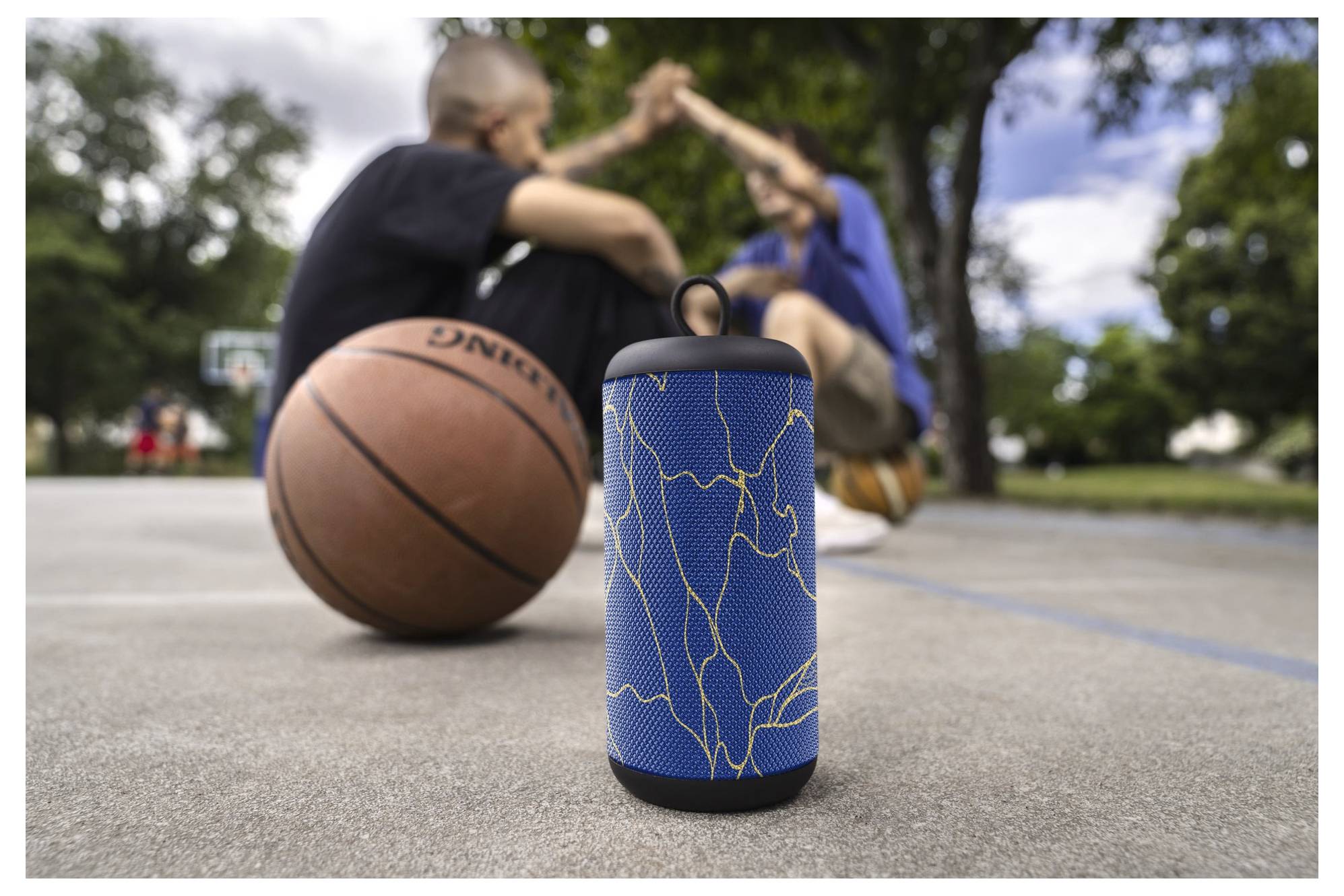 Ein tragbarer Lautsprecher mit blauem Design sitzt auf einem Basketballplatz in der Nähe eines Basketballs, mit zwei unscharfen Personen im Hintergrund.