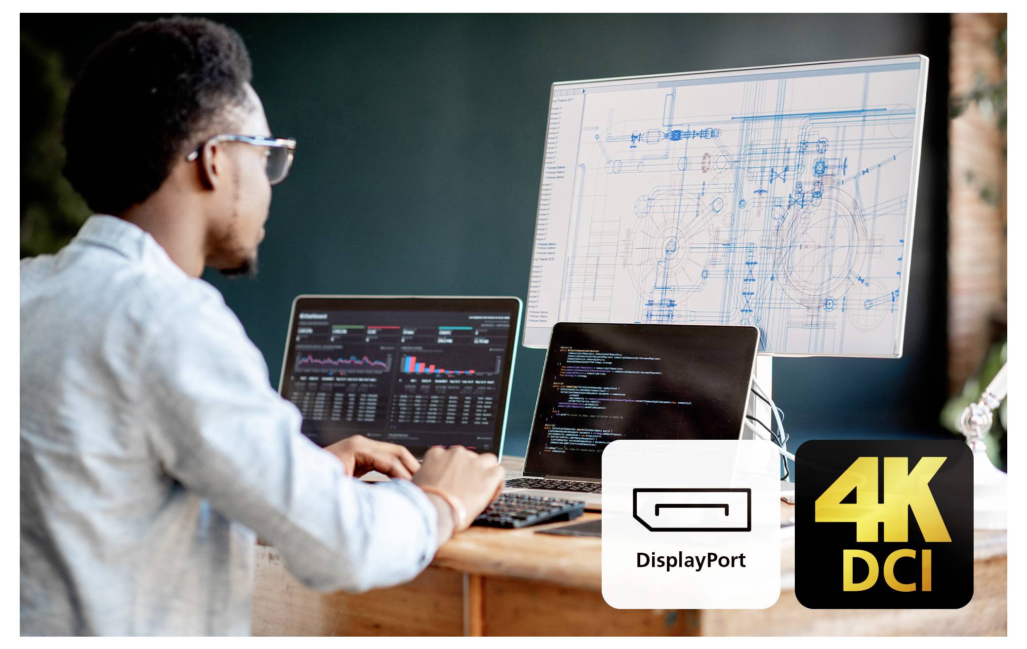 Eine Person, die an einem Schreibtisch mit zwei Laptops und einem Monitor arbeitet, der technische Schemazeichnungen und Daten zeigt, wobei 'DisplayPort' und '4K DCI' hervorgehoben sind.