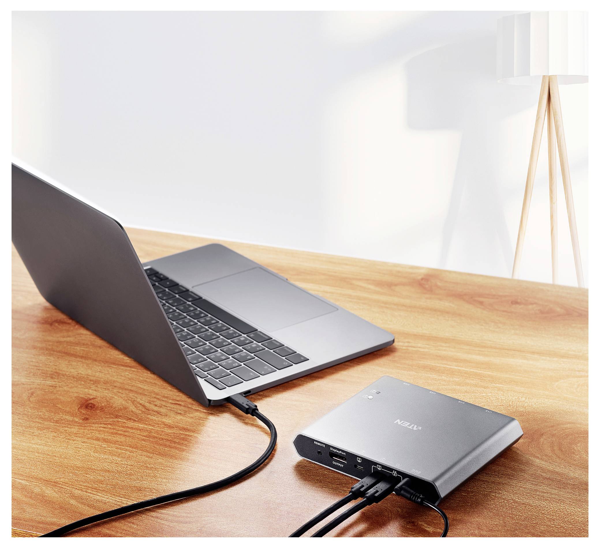 Ein silberner Laptop, der mit einer grauen Dockingstation auf einem Holzschreibtisch verbunden ist, mit Kabeln, die zu anderen Geräten führen, unter einer modernen Lampe.