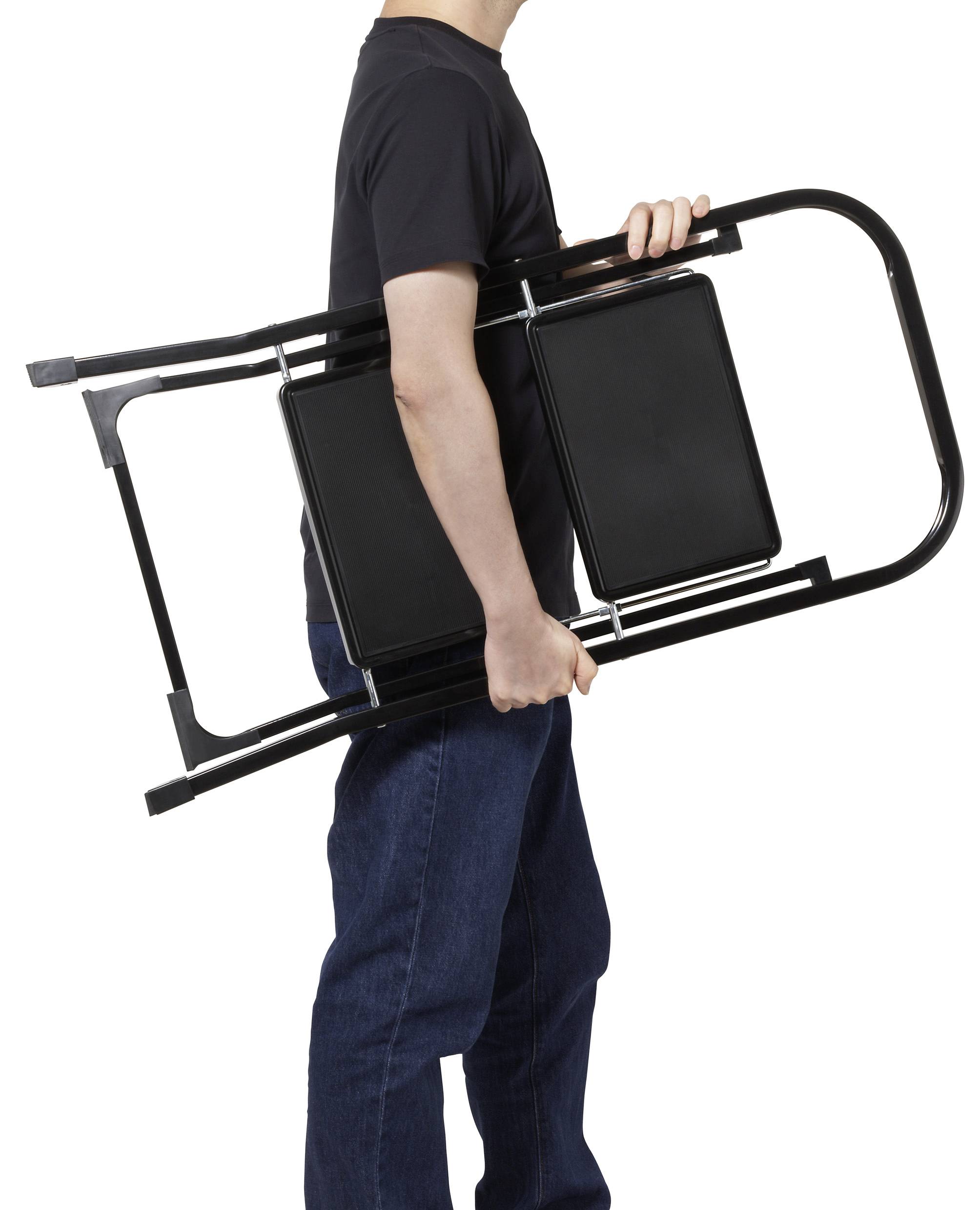 Eine Person in einem schwarzen T-Shirt und blauen Jeans trägt einen zusammengefalteten schwarzen Metallstuhl.